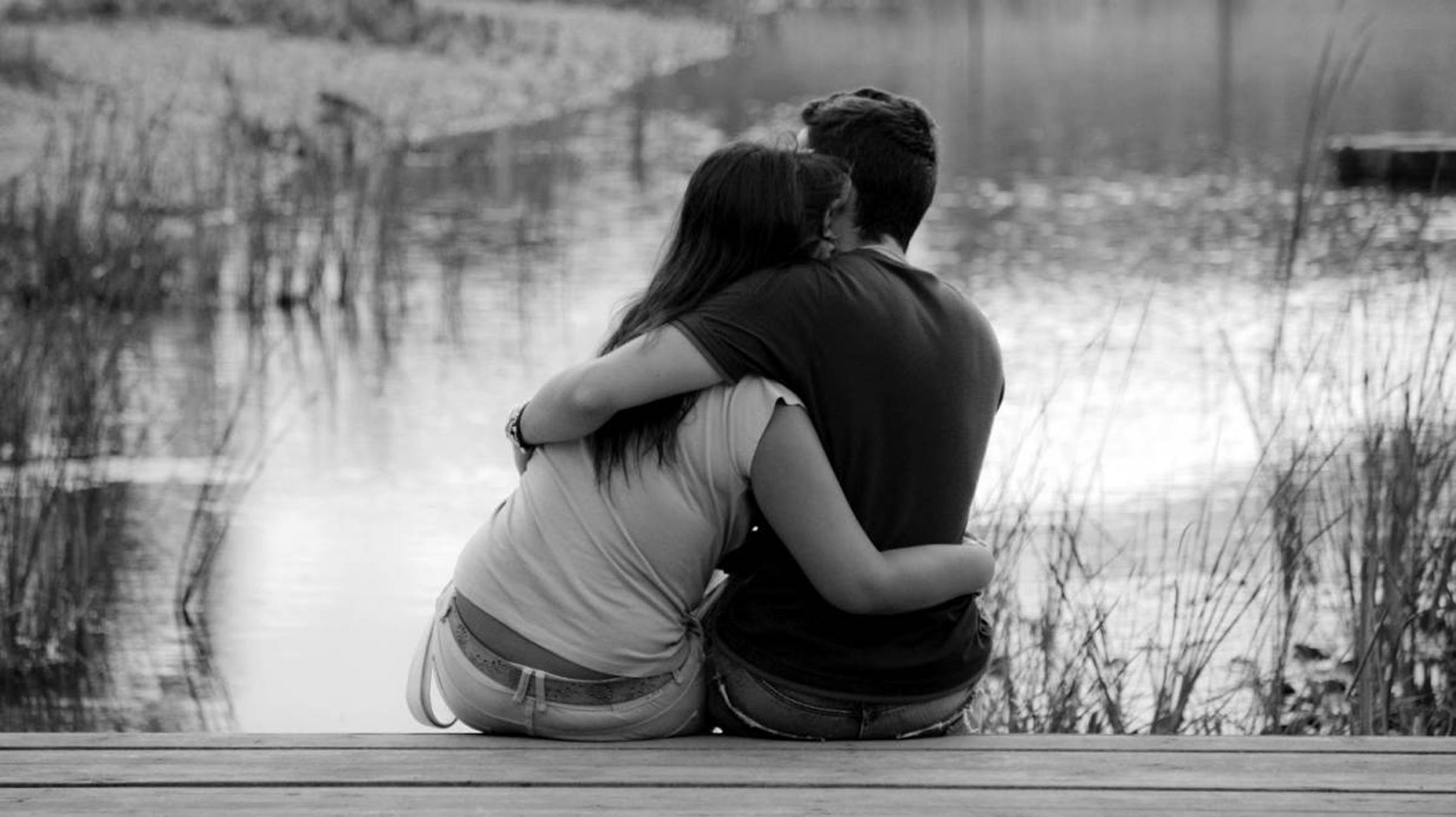Pareja de novios jóvenes de espalda, abrazándose sentados en un muelle frente a un lago