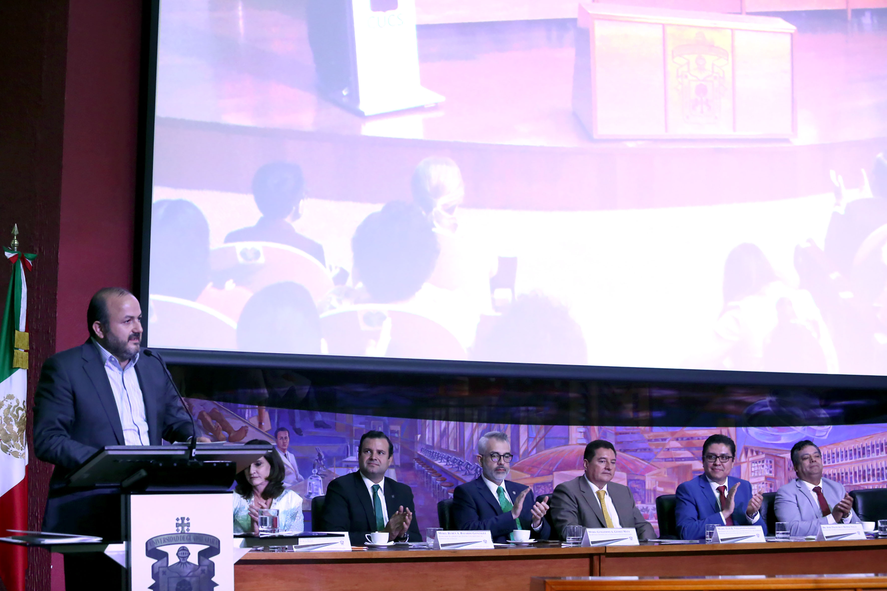 Rector General al micrófono en pódium dando mensaje tras informe, al fondo miembros del presídium