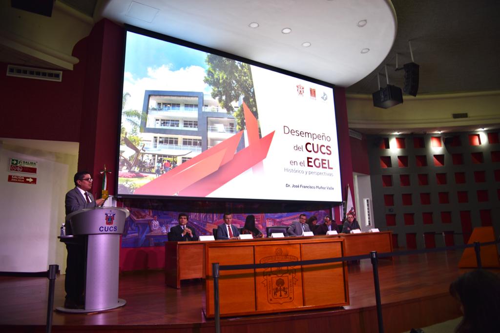 Rector del CUCS al micrófono en el pódium, toma abierta, al fondo una diapositiva en la pantalla y los miembros del presídium