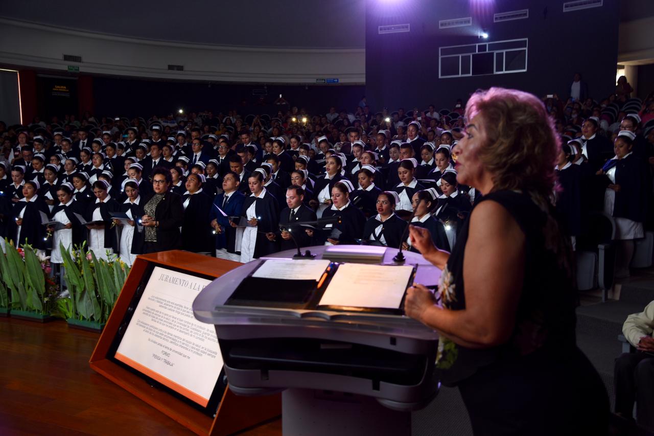 Coordinadora de carrera ofreciendo mensaje a graduados en el pódium, al fondo el auditorio lleno a toda su capacidad