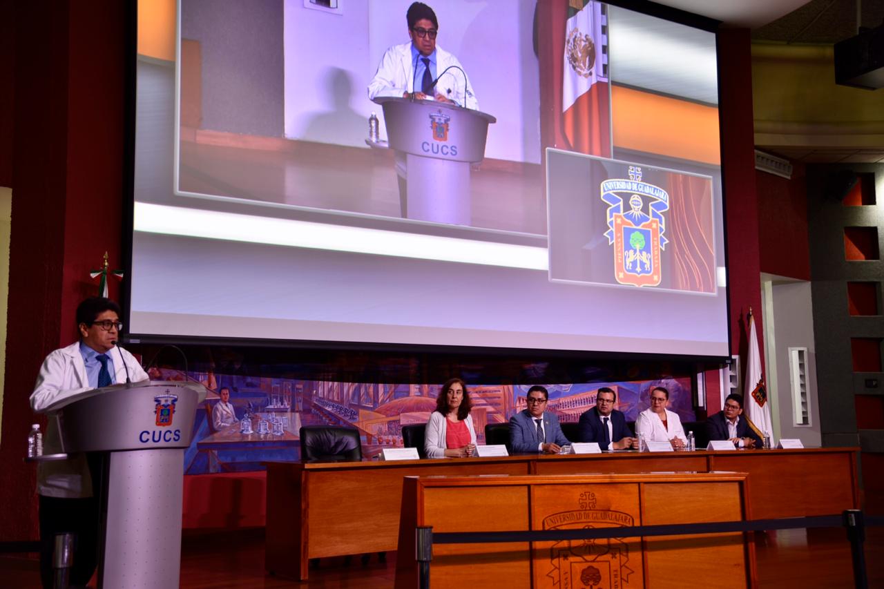 Coordinador de Investigación del IMSS al micrófono en el pódium, al fondo los miembros del presídium y en la pantalla su imagen