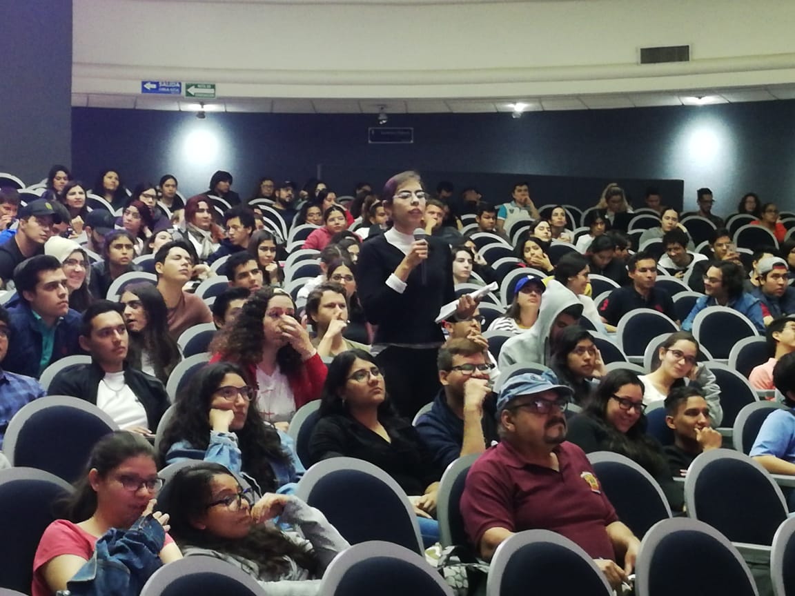 Vista general de los asistentes a la conferencia. Una alumna de pie tomando la palabra