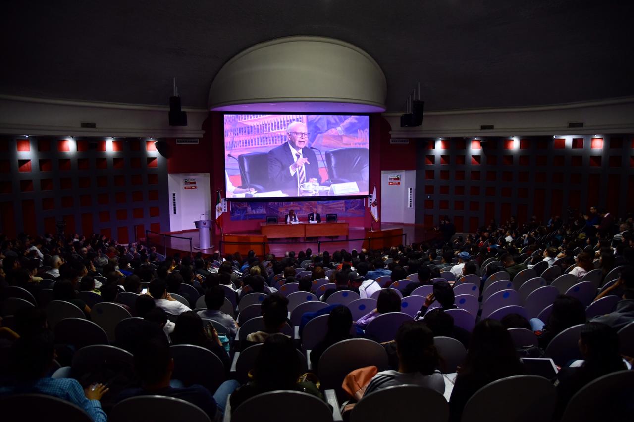 Toma abierta desde atrás del auditorio que luce lleno, al fondo en la pantalla la imagen del conferencista