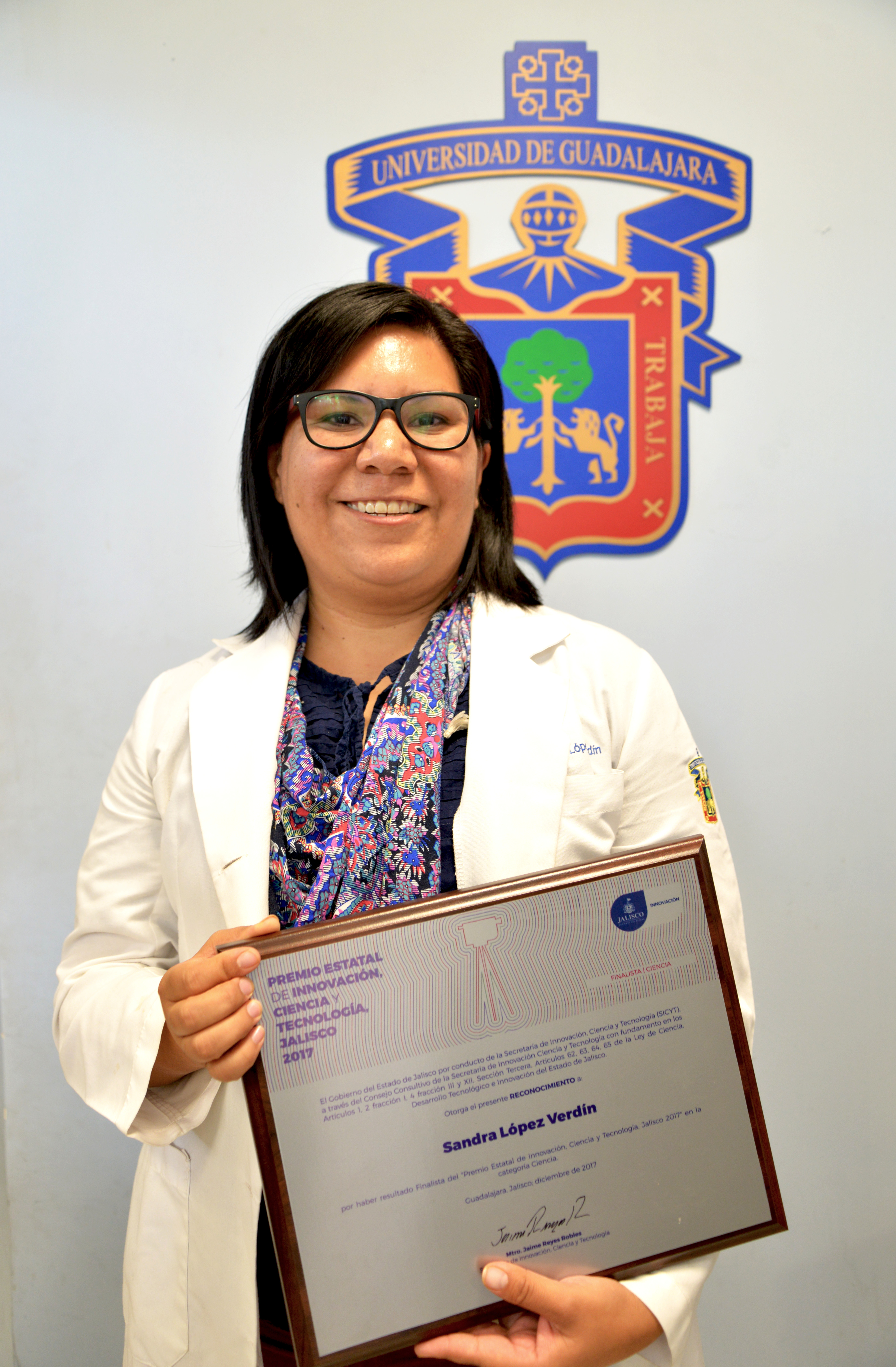 Dra. Silvia López Verdín exhibiendo su reconocimiento, atrás el escudo de la UdeG