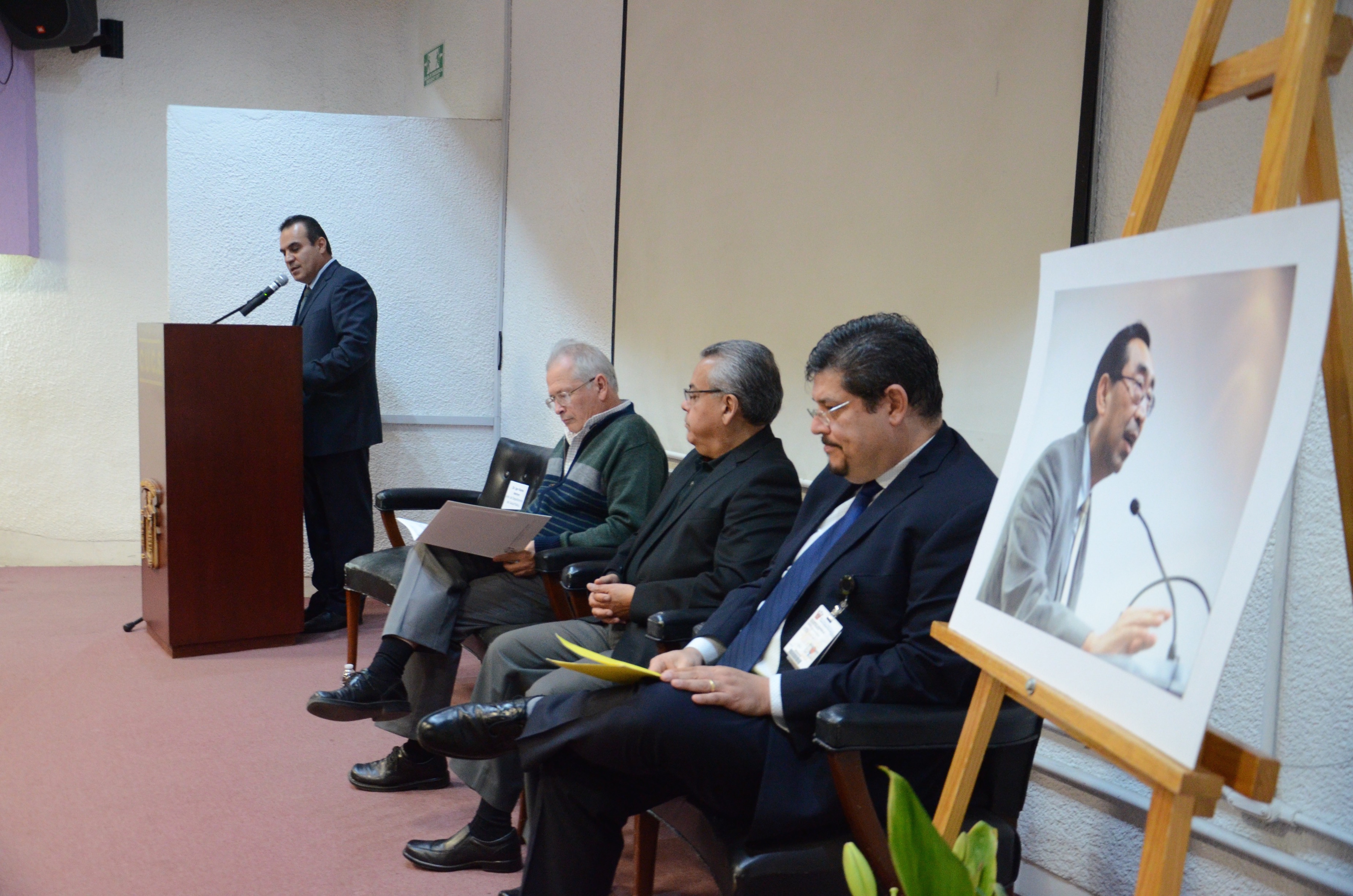 En primer plano fotografía del Dr. Roberto Kumasawa, atrás miembros del presídium