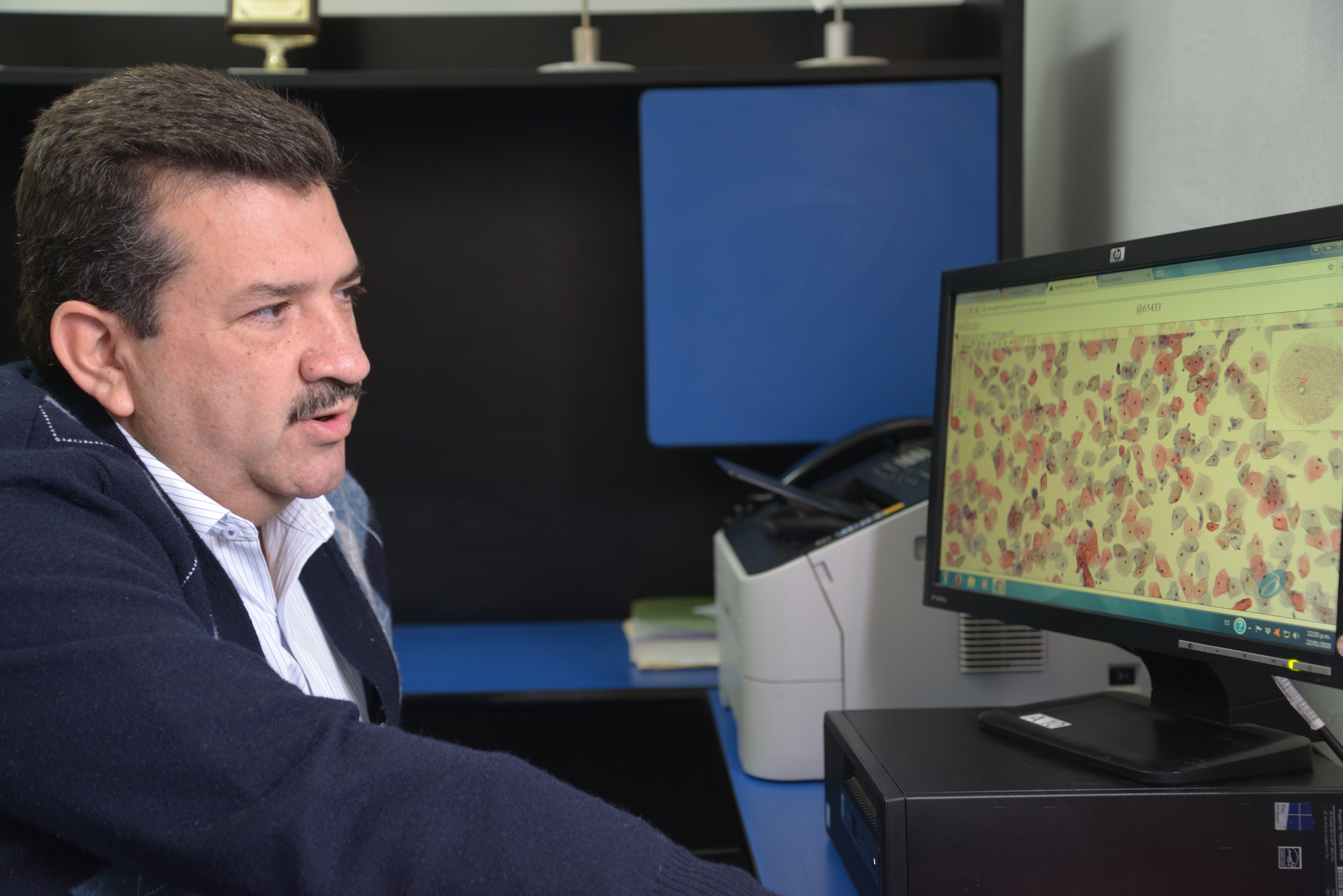 Dr. Bañuelos en entrevista, al fondo la pantalla de su computadora muestra un corte histológico visto con el microscopio virtual