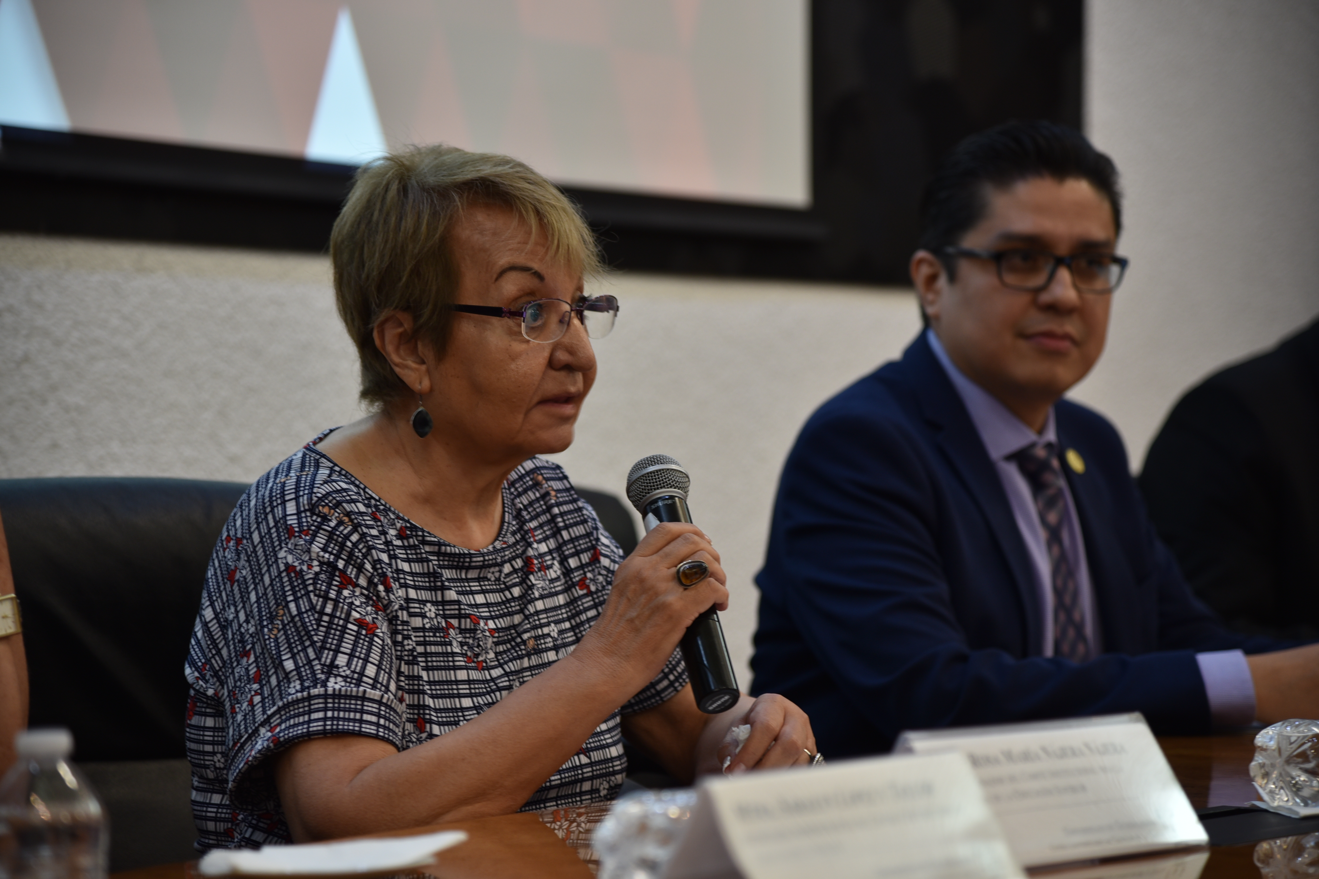 Coordinadora de pares académicos al micrófono en la mesa del presídium, a su lado el rector del CUCS