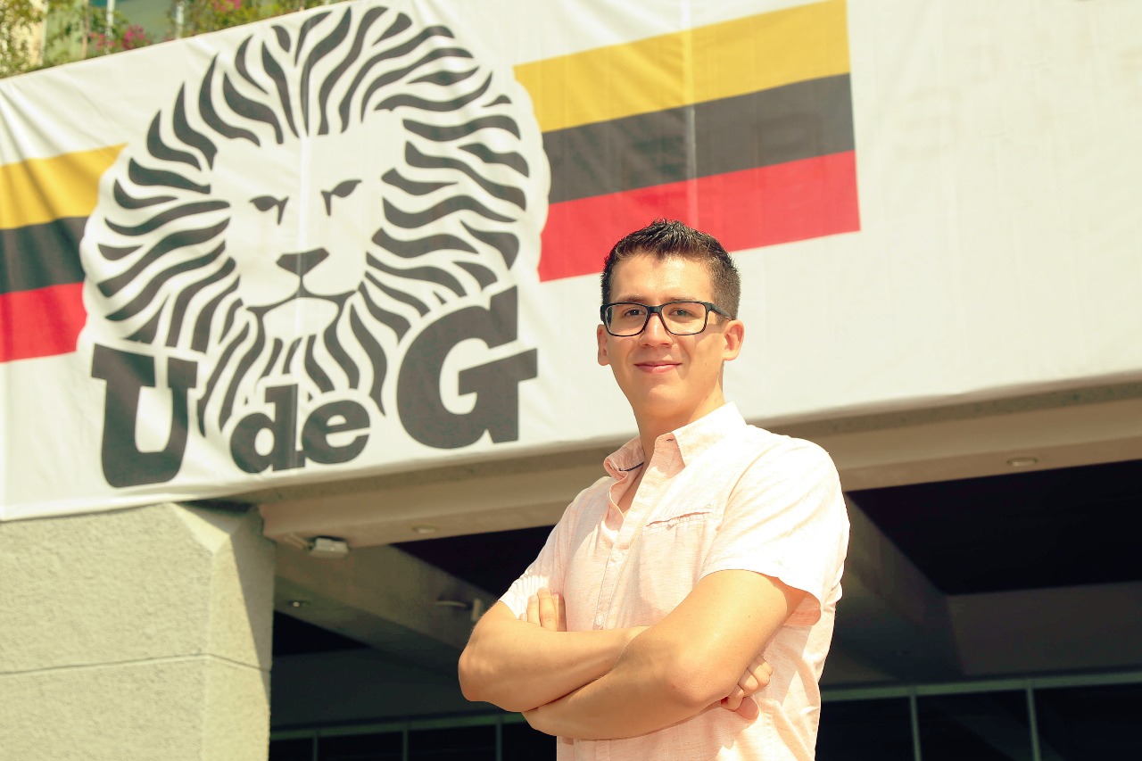 Alumno posa en la explanada del Edificio de Rectoría General, a sus espaldas el escudo de la UdeG